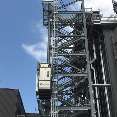 ONEMcDonough A metal industrial elevator shaft and stairway structure stands against a blue sky, with pipes and buildings in the background—an ideal setting to compare Rack and Pinion vs. Traction Elevators in demanding environments.