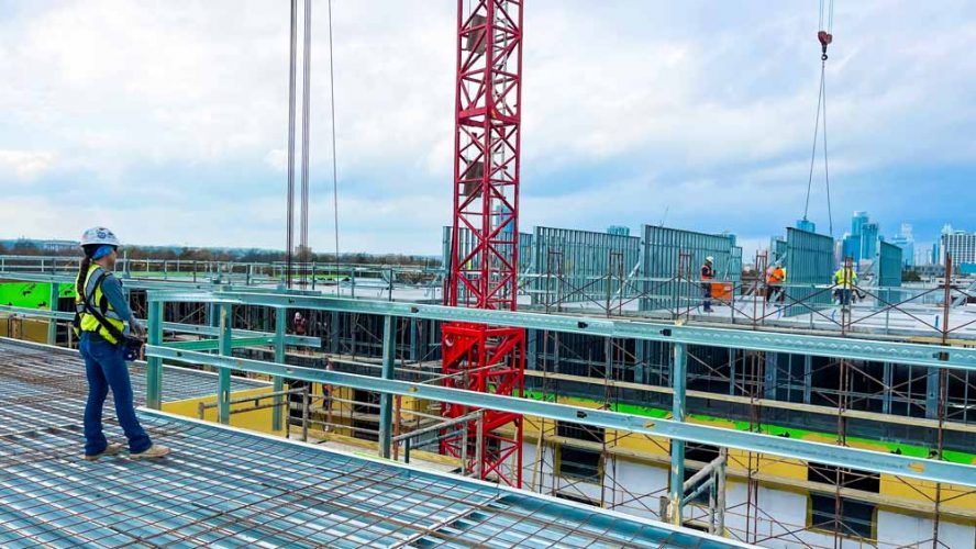 ONEMcDonough Construction workers on a building site with scaffolding, a red crane, ONEMcDonough Elevators and Rentals equipment, and steel framework under a cloudy sky.
