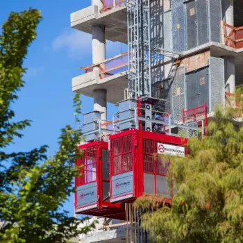 ONEMcDonough A red construction hoist rental is attached to the side of a partially built concrete building, with trees in the foreground and a clear sky in the background.