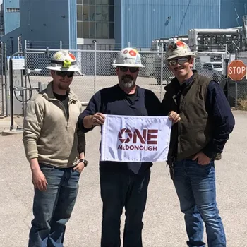 ONEMcDonough Three men in work attire and helmets stand outside an industrial facility, proudly holding a small flag that reads One McDonough.