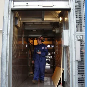 ONEMcDonough A person in a blue coverall and hard hat walks through a metal industrial hallway, holding equipment and looking to the side, performing elevator service and maintenance.