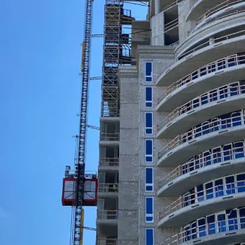 ONEMcDonough A construction elevator and scaffolding, with construction hoist rental equipment, are attached to the side of a high-rise building under construction against a clear blue sky.