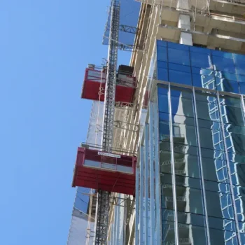 ONEMcDonough Construction hoist rental units are attached to the side of a tall building with reflective glass windows under a clear blue sky.