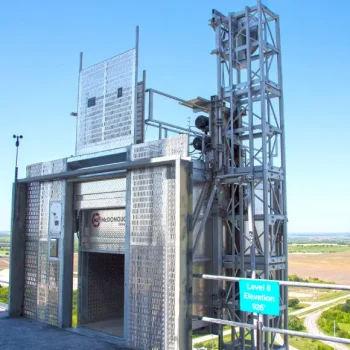 ONEMcDonough A metal construction hoist elevator stands on an outdoor platform at level 8, elevation 926, overlooking open fields and roads under a clear blue sky—ideal for a construction elevator consultation.