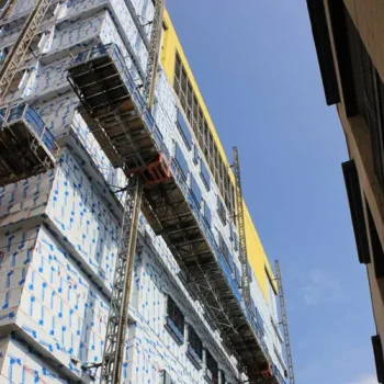 ONEMcDonough A multi-story building under construction with scaffolding, exterior insulation panels, and a McDonough Elevator, viewed from below against a blue sky.