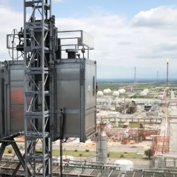 ONEMcDonough Rack and Pinion Elevators are featured in this industrial construction elevator, attached to a metal tower overlooking a large refinery complex with storage tanks and extensive infrastructure in the background.