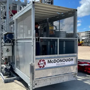 ONEMcDonough A metal construction elevator with a McDonough Elevators sign, available through construction equipment rentals, is stationed outdoors on a sunny day with industrial buildings in the background.